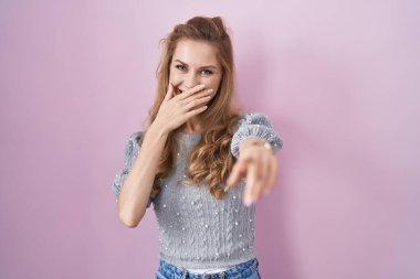 Güzel sarışın kadın pembe arka planda durup sana gülüyor, eli ağzında kamerayı işaret ediyor, yüz ifadesi utanç verici. 