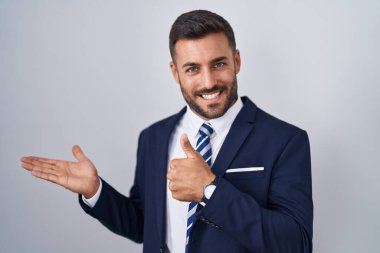 Yakışıklı İspanyol takım elbiseli ve kravatlı adam avuç içi gösteriyor ve baş parmaklarıyla iyi bir jest yapıyor, mutlu ve neşeli gülümsüyor. 