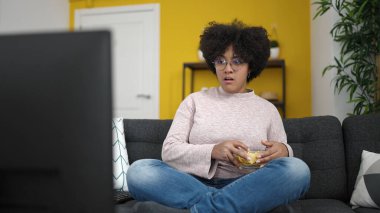Genç Afrikalı Amerikalı kadın televizyon seyrediyor. Kanepede oturup cips yiyor.