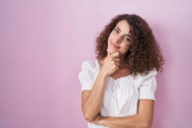 Hispanic woman with curly hair standing over pink background looking confident at the camera smiling with crossed arms and hand raised on chin. thinking positive. 