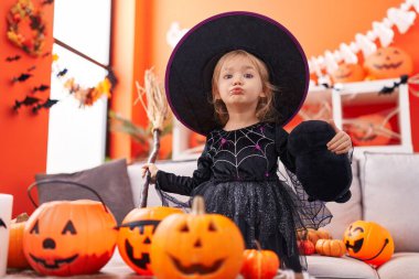 Cadı kostümü giyen sevimli sarışın kız evde süpürge tutuyor.