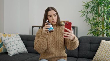 Genç ve güzel İspanyol bir kadın evde elinde ilaç şişesiyle çevrimiçi tıbbi danışmanlık yapıyor.