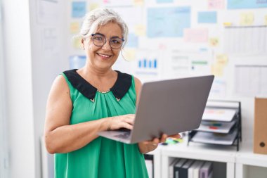 Ofiste çalışırken dizüstü bilgisayar kullanan, orta yaşlı, kır saçlı iş kadını.
