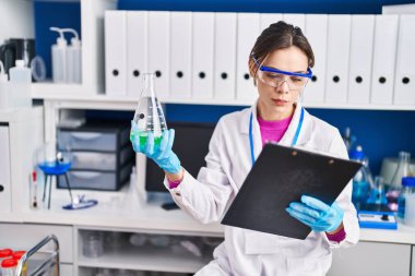 Young beautiful hispanic woman scientist reading report holding test tube at laboratory