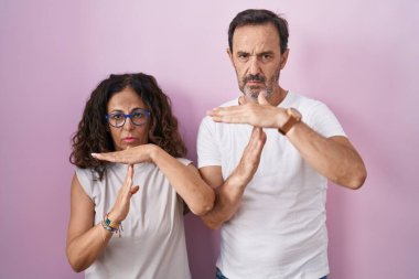 Orta yaşlı İspanyol çift pembe arka planda el kol hareketleri yapıyor, hayal kırıklığına uğramış ve ciddi bir yüz ifadesi takınıyor. 
