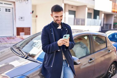 Young hispanic man using smartphone leaning on car at street