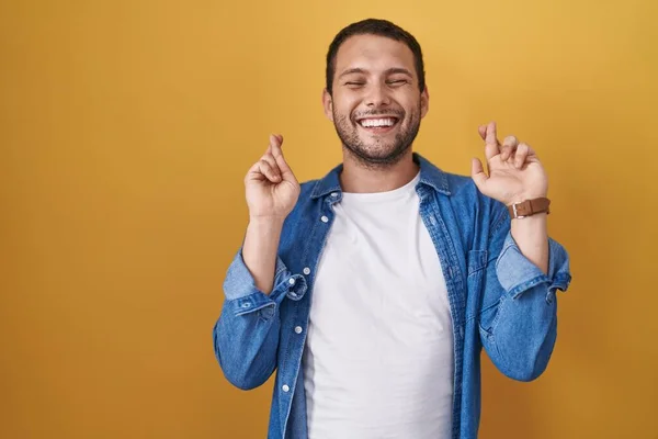黄色い背景のジェスチャー指の上に立つヒスパニック系の男性は希望と目を閉じて笑顔を渡った 幸運と迷信の概念は — ストック写真