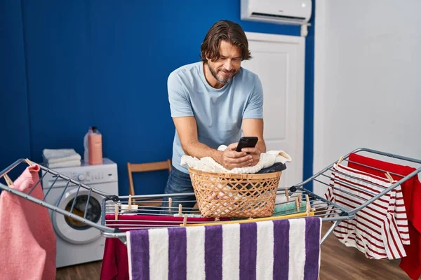 中年男子在洗衣房用智能手机把衣服挂在晾衣绳上 — 图库照片