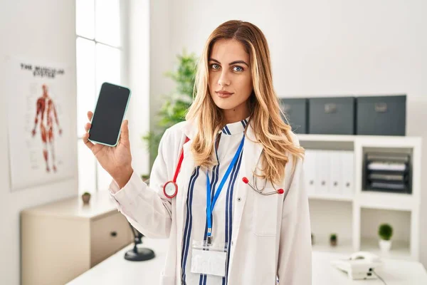 Young Blonde Doctor Woman Working Clinic Showing Smartphone Screen Thinking — Stock Photo, Image