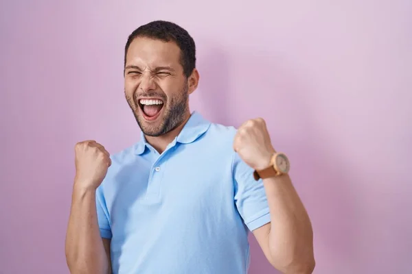 ピンクの背景に立っているヒスパニック系の男は非常に幸せと腕を上げて勝者のジェスチャーを行う興奮 笑顔と成功のために叫んで お祝いのコンセプト — ストック写真