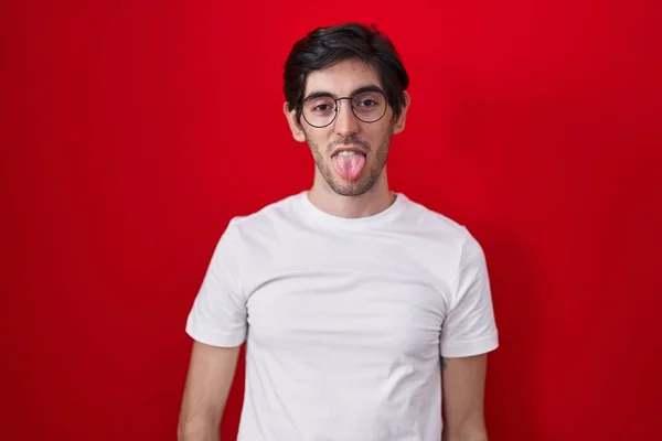 Mladý Hispánec Který Stojí Nad Červeným Pozadím Radostně Vystrkuje Jazyk — Stock fotografie