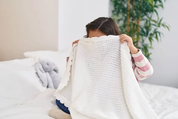 stock image Adorable hispanic girl sitting on bed covering face with blanket at bedroom