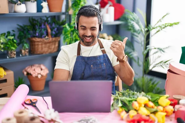 Hispanic man with beard working at florist shop using laptop pointing thumb up to the side smiling happy with open mouth