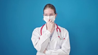 Sarışın genç kadın doktor maskesini çıkarıyor mavi arka planda gülümsüyor.