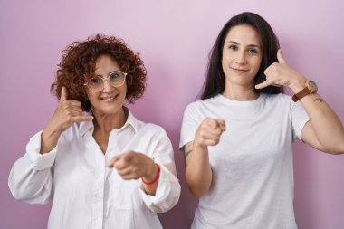 İspanyol anne ve kızı pembe arka plan üzerine günlük beyaz t-shirt giyip gülümsüyor telefon jesti hakkında konuşuyor ve seni işaret ediyorlar. Ara beni.. 
