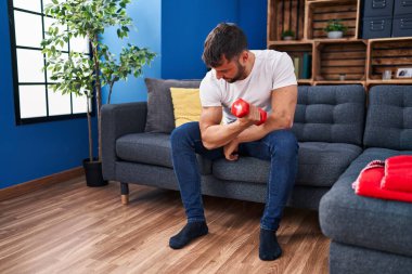 Young hispanic man training using dumbbell sitting on sofa at home