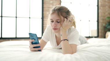 Yatak odasında uzanmış akıllı telefon kullanan genç sarışın kadın.