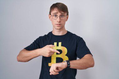 Bitcoin tişörtü giyen beyaz tenli sarışın adam acele ederek saati, sabırsızlığı ve gecikmeye kızmayı işaret ediyor. 