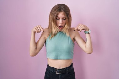 Blonde caucasian woman standing over pink background pointing down with fingers showing advertisement, surprised face and open mouth 