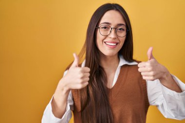 Sarı arka planda duran esmer, gözlüklü, başarı işareti takan, el hareketi yapan, gülümseyen ve mutlu genç bir kadın. Neşeli ifade ve kazanan jest. 
