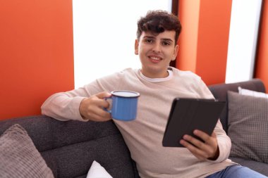Non binary man using touchpad drinking coffee sitting on sofa at home