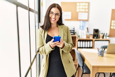 Genç, güzel İspanyol iş kadını ofisteki akıllı telefonu kullanarak gülümsüyor.