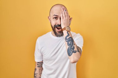 Sarı arka planda duran İspanyol dövmeli genç bir adam bir gözünü eliyle kapatıyor, yüzünde kendine güvenen bir gülümseme ve şaşırtıcı bir duygu var.. 