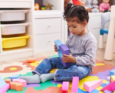 Adorable hispanic girl playing with construction blocks sitting on floor at kindergarten