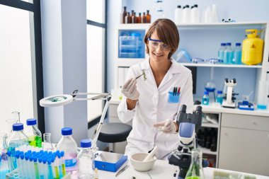 Young beautiful hispanic woman scientist smiling confident holding plant with tweezer at laboratory