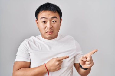 Beyaz arka planda duran genç Çinli bir adam iki eli de endişeli ve gergin, endişeli ve şaşırmış bir ifadeye işaret ediyor. 