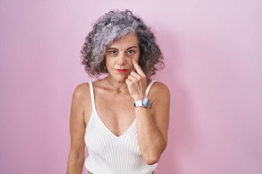 Middle age woman with grey hair standing over pink background pointing to the eye watching you gesture, suspicious expression 