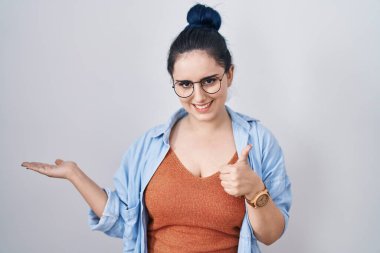 Beyaz arka planda duran mavi saçlı genç modern kız avuç içi elini gösteriyor ve baş parmaklarıyla iyi bir hareket yapıyor, mutlu ve neşeli gülümsüyor. 