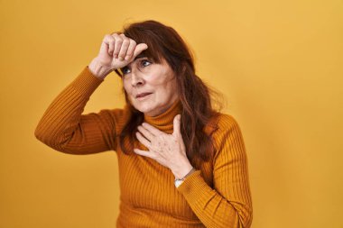 Orta yaşlı İspanyol kadın sarı arka planda duruyor. Hastalık, ateş, grip ve virüs için alnına dokunuyor. 