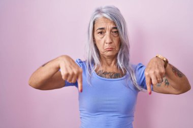 Pembe arka planda duran orta yaşlı kadın üzgün ve üzgün görünüyor, parmaklarıyla yön gösteriyor, mutsuz ve depresif.. 