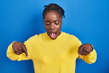 Beautiful black woman standing over blue background pointing down with fingers showing advertisement, surprised face and open mouth 