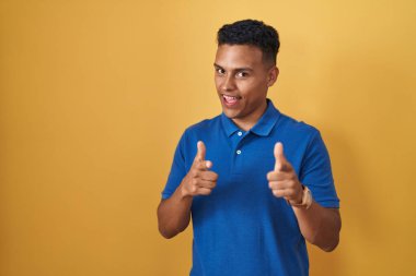 Young hispanic man standing over yellow background pointing fingers to camera with happy and funny face. good energy and vibes. 