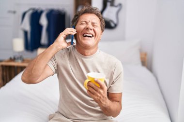 Orta yaşlı bir adam akıllı telefondan konuşuyor. Yatak odasında kahve içiyor.