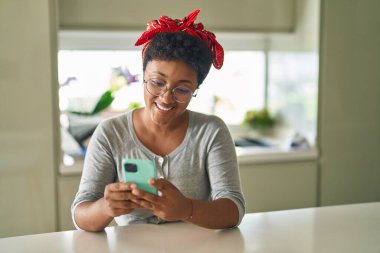 Afro-Amerikalı kadın akıllı telefon kullanıyor. Evde masada oturuyor.