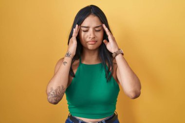 Sarı arka planda duran esmer kadın, elleri başının üstünde, baş ağrısı var çünkü stres. migren ağrısı. 