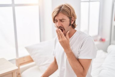 Young blond man sitting on bed yawning at bedroom