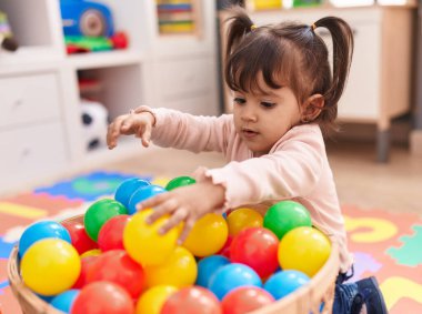 Adorable hispanic girl playing with balls sitting on floor at kindergarten