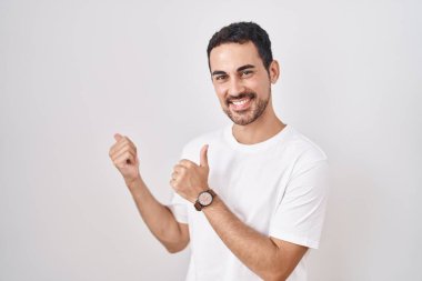 Beyaz arka planda duran yakışıklı İspanyol adam. Arkayı işaret ediyor. Elleri havada, kendinden emin gülümsüyor. 