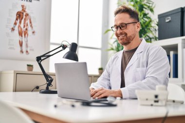 Klinikte çalışan genç beyaz bir doktor dizüstü bilgisayar kullanıyor.