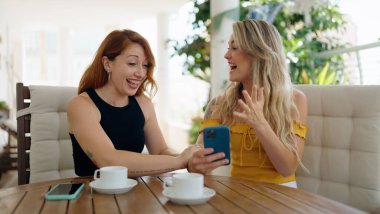 Akıllı telefon kullanan ve kahve içen iki kadın evdeki terasta oturuyor.