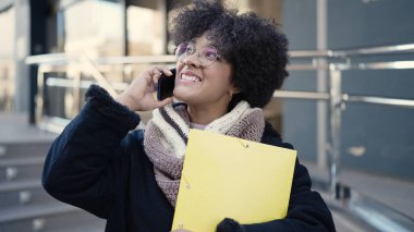 Genç Afrikalı Amerikalı kadın gülümsüyor. Elinde klasörle caddede telefonla konuşuyor.