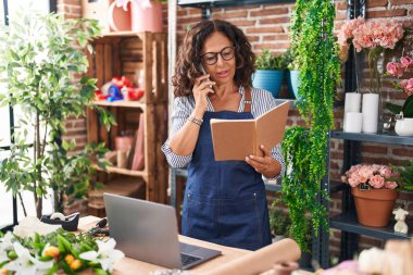 Middle age woman florist talking on smartphone reading book at flower shop