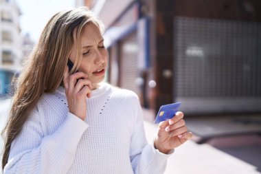 Genç sarışın kadın elinde kredi kartıyla akıllı telefondan konuşuyor.