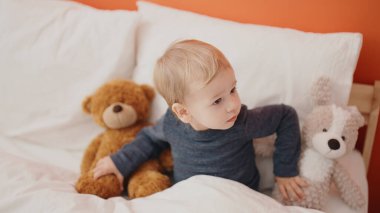 Sevimli sarışın bebek yatak odasında oyuncak ayıyla yatakta oturuyor.