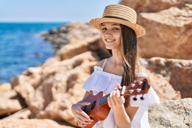 Sevimli kız turist, deniz kenarında ukulele oynarken kendine güvenerek gülümsüyor.