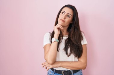 Pembe arka planda eli çenesinde duran genç esmer kadın soru sormayı, düşünceli yüz ifadesini düşünüyor. Düşünceli bir yüzle gülümsüyor. şüphe kavramı. 